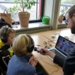 Zwei Kinder hören mit Kopfhörern zu, während ein Erwachsener spricht und ein Laptop bedient.