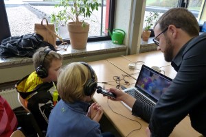 Zwei Kinder hören mit Kopfhörern zu, während ein Erwachsener spricht und ein Laptop bedient.