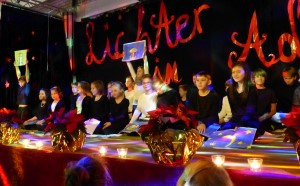 Festliche Feier mit Kindern auf der Bühne, umgeben von Weihnachtsdeko und Kerzenlicht.