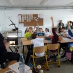 Lehrer spricht zu Schülern in einem Klassenzimmer; viele Hände sind oben.