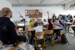 Lehrer spricht zu Schülern in einem Klassenzimmer; viele Hände sind oben.