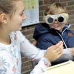 Ein Kind mit speziellen Brillen sitzt am Tisch und interagiert mit einer älteren Person.