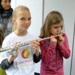 Mädchen spielt Flöte, andere Kinder hören zu, eine Person im Hintergrund.