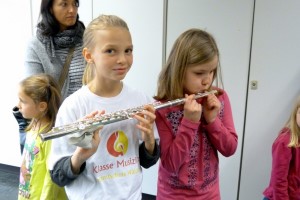 Mädchen spielt Flöte, andere Kinder hören zu, eine Person im Hintergrund.