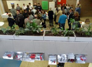Menschen stehen in einer Aula, im Vordergrund Pflanzen und kleine Objekte in Kästen.
