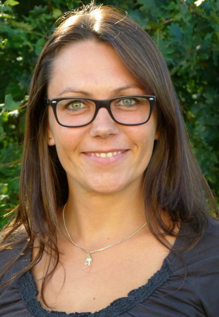 Frau mit langen Haaren und Brille lächelt vor einem grünen Hintergrund.