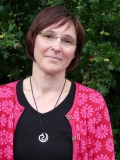 Eine Person mit schulterlangen Haaren trägt einen pinken Pullover und steht vor einem grünen Hintergrund.