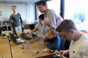 Gruppe von Schülern arbeitet an Projekten an einem Tisch mit Computern und Materialien.