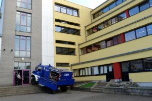 Ein blauer Fahrzeug steht schräg vor einem Schulgebäude mit mehreren Etagen.