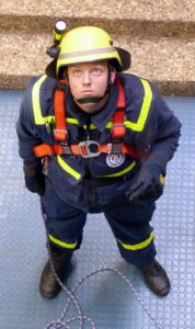 Feuerwehrmann in Schutzkleidung schaut nach oben, mit Sicherheitsgurt und Seil.