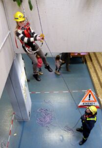 Person in Kletterausrüstung schwebt an Seil in einem Gebäude. Feuerwehrmann steht daneben.