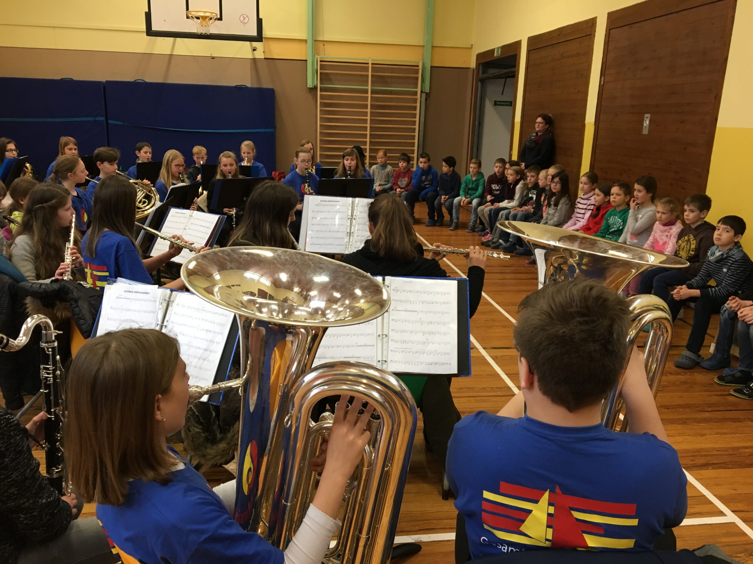 Schüler*innen im Musikunterricht spielen Instrumente, Publikum sitzt auf Bänken.