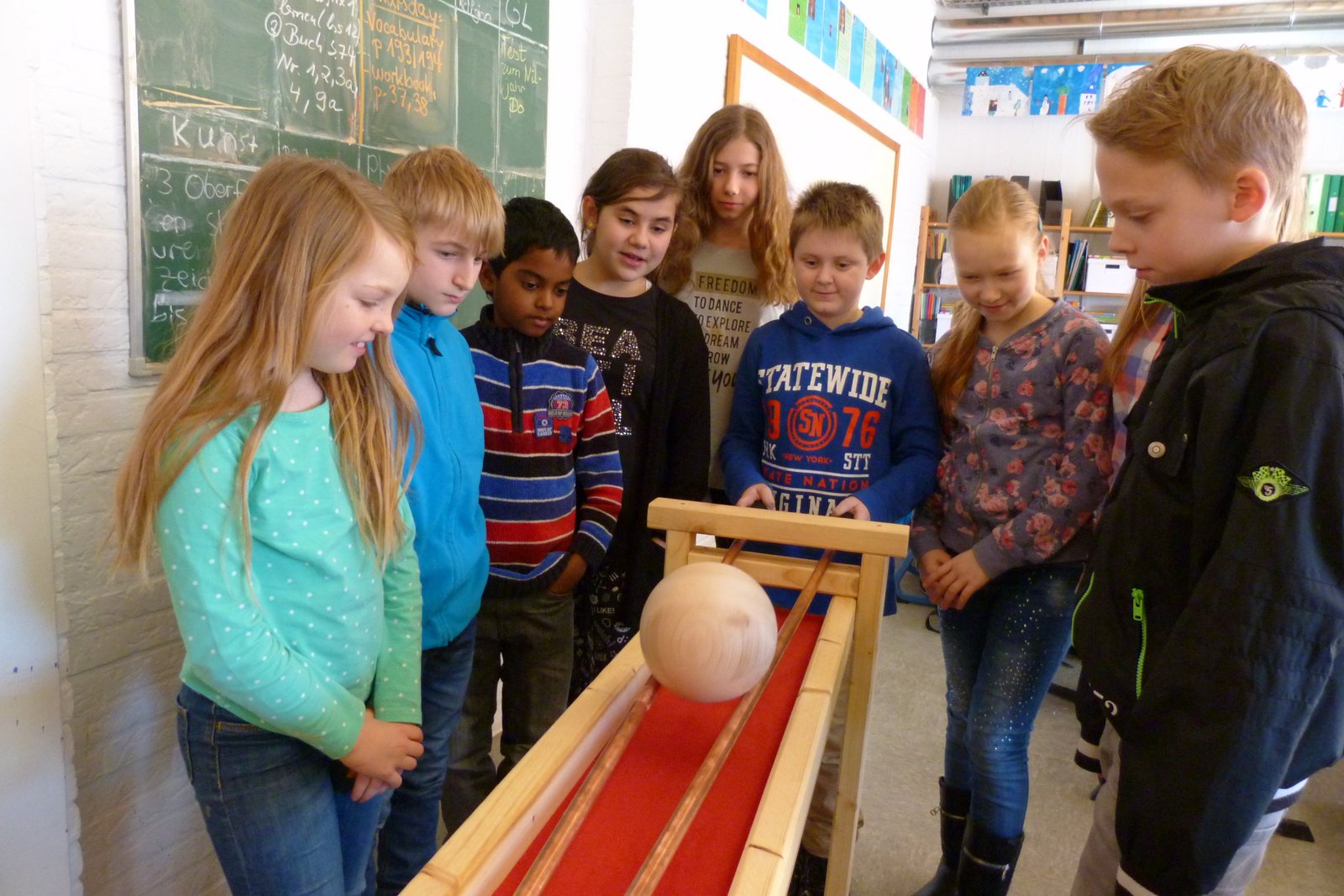 Schülerinnen und Schüler beobachten gespannt, wie eine Holzkugel eine schiefe Ebene hinunterrollt – ein physikalisches Experiment im Klassenraum.