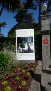 Ein großes Informationsschild über Christian Dior in Granville, umgeben von Blumen und Pflanzen.