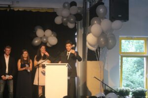 Schüler steht am Rednerpult, Gruppe im Hintergrund, Dekoration mit Schwarz-Weiß-Luftballons.