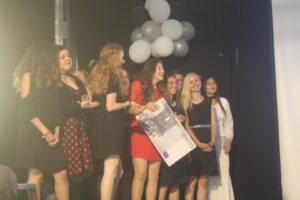 Gruppe von Frauen in festlicher Kleidung auf einer Bühne mit Luftballons.