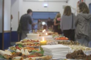 Buffet mit verschiedenen Speisen und Getränken in einem Raum, Gäste im Hintergrund.