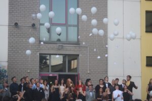 Abiturienten werfen weiße Luftballons vor einer Schule in die Luft.