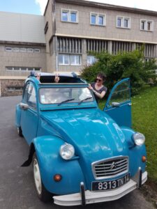 Blauer Citroën 2CV mit offener Tür und einer Person daneben. Grünes Gras im Hintergrund.