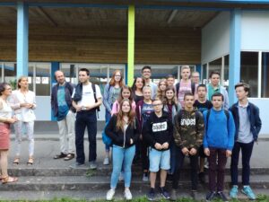 Gruppe von Schülern und Lehrern vor einem Schulgebäude, einige stehen, andere sitzen.