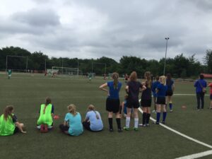 Mädchen sitzen am Spielfeldrand und beobachten ein Fußballspiel.