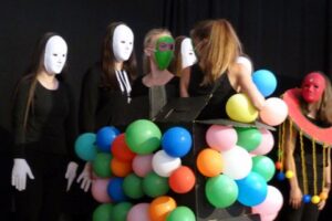 Schülerinnen in Kostümen mit Masken und bunten Ballons auf der Bühne.