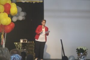 Frau in rotem Blazer spricht auf einer Bühne vor Luftballons.