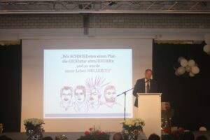 Ein Redner steht am Pult vor einer Präsentation mit bunten Figuren und Text.