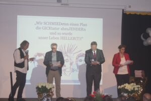 Vier Personen stehen auf einer Bühne und lesen Texte vor. Im Hintergrund sieht man eine Präsentation.
