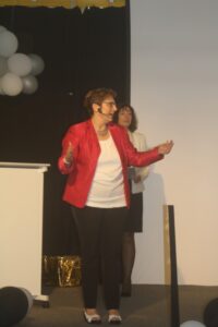 Eine Frau in rotem Blazer spricht auf einer Bühne, im Hintergrund steht eine weitere Frau.