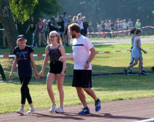 Mädchen und ein Mann gehen Hand in Hand auf einer Sportbahn, Zuschauer im Hintergrund.