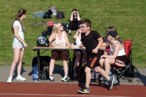 Schülerinnen und Schüler sitzen und stehen auf einem Sportplatz, draußen bei Sonnenschein.
