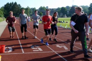 Schüler joggen auf der Laufbahn, andere stehen am Rand. Sportveranstaltung im Freien.