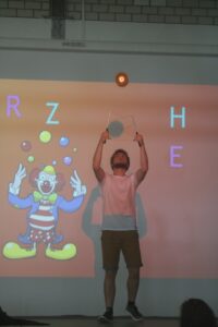 Ein Mann jongliert mit einem Ball und einer Platte vor einer Projektion eines Clowns.