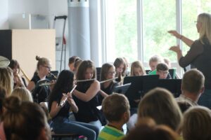 Schüler*innen spielen im Orchester, eine Lehrkraft dirigiert. Publikum sitzt im Vordergrund.