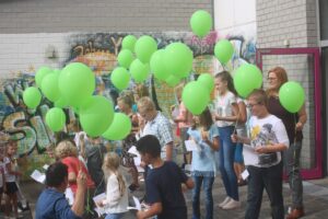 Schüler halten grüne Ballons und feiern vor einer bunten Wand.