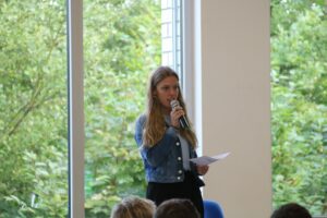 Schülerin spricht mit Mikrofon vor einem Publikum und Fensterblick ins Grüne.