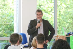 Ein Mann spricht mit einem Mikrofon vor einem Publikum in einem hellen Raum.