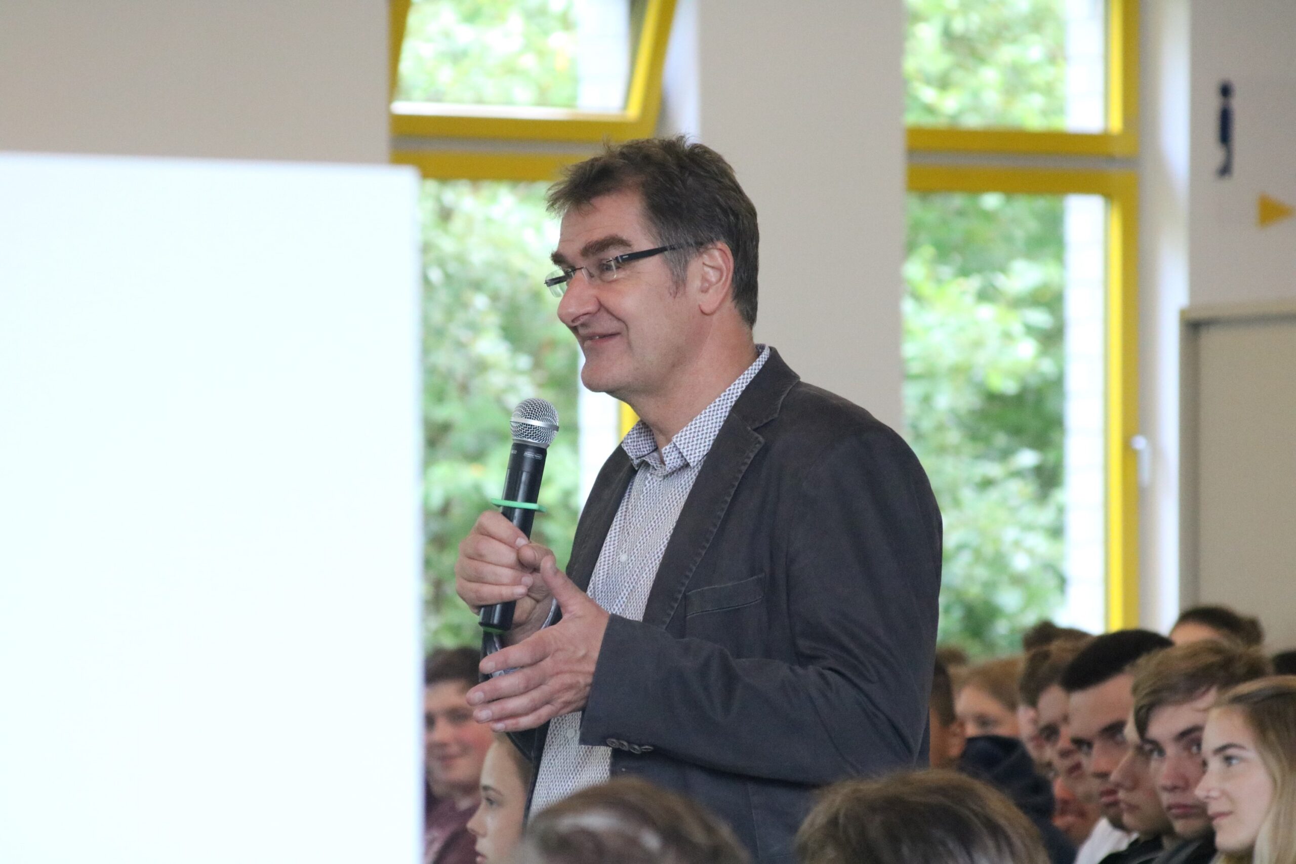 Mann spricht mit Mikrofon vor einer Gruppe von Schülern in einem Klassenzimmer.