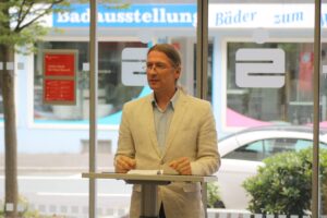 Mann steht an einem Tisch und spricht in einem modernen Raum mit großer Fensterfront.