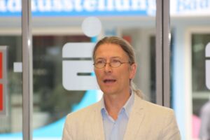 Mann mit Brille und langem Haar spricht vor Glaswand.