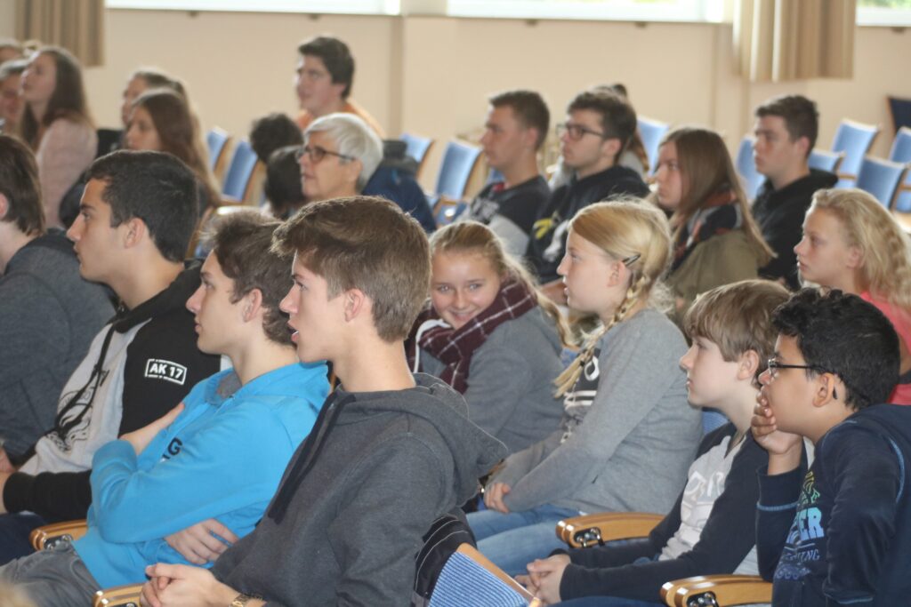 Schüler und Erwachsene sitzen in einem Hörsaal und hören aufmerksam zu.