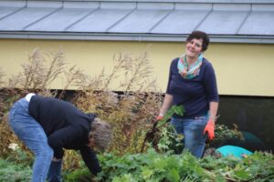 Zwei Frauen pflücken Pflanzen im Garten, umgeben von grünem Bewuchs.
