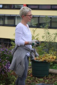 Frau mit Brille und kurzem Haar steht im Garten mit Pflanzen.