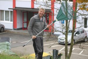 Mann mit Rechen steht vor einem Gebäude und lächelt.