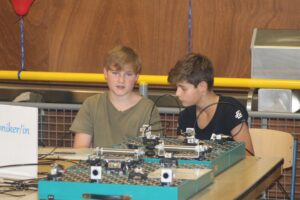 Zwei Jungen sitzen an einem Tisch mit technischen Geräten und diskutieren.