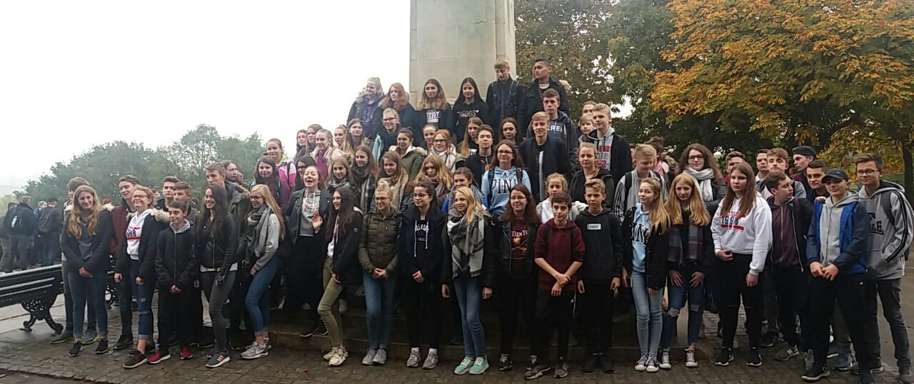 Gruppe von Schülern posiert vor einem Denkmal im Freien an einem bewölkten Tag.
