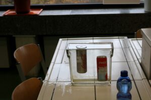 Zwei Getränkedosen und eine Flasche Wasser in transparentem Behälter auf einem Tisch.