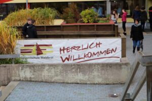 Banner mit der Aufschrift "Herzlich Willkommen" an einer Schule, umgeben von Pflanzen.
