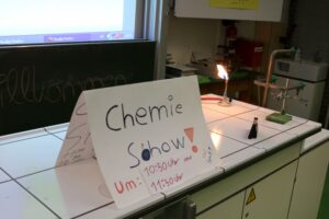 Chemietisch mit Plakat zur Chemieshow und einer brennenden Flamme im Hintergrund.
